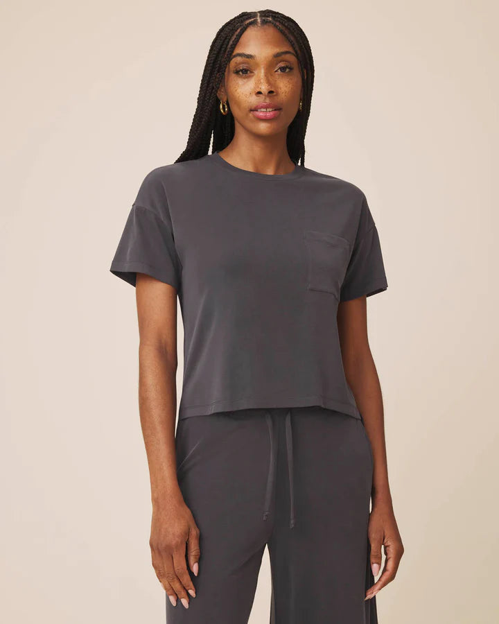 A woman modeling a dark gray short-sleeve crop top with a chest pocket, paired with matching dark gray drawstring pants, against a solid beige background.