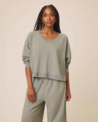 A woman modeling a matching sage green lounge set consisting of a cropped long-sleeve V-neck top and coordinating wide-leg pants, set against a plain beige studio background.