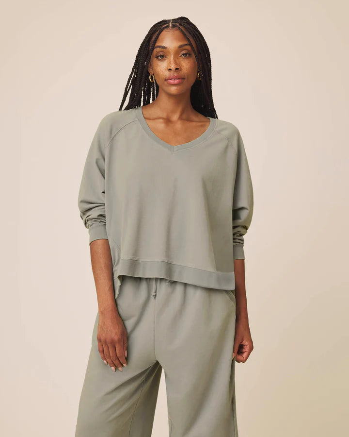 A woman modeling a matching sage green lounge set consisting of a cropped long-sleeve V-neck top and coordinating wide-leg pants, set against a plain beige studio background.