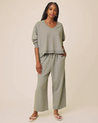 A woman modeling a matching sage green lounge set consisting of a cropped long-sleeve V-neck top and wide-leg cropped pants, paired with white flip-flop sandals against a solid beige studio background.