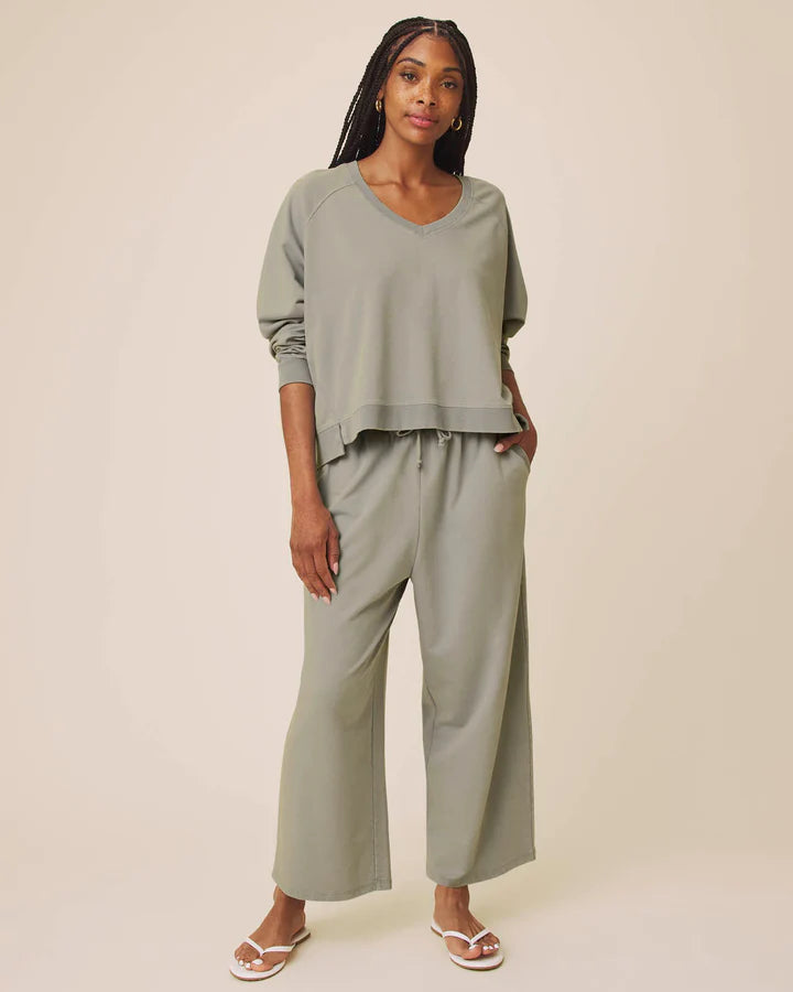 A woman modeling a matching sage green lounge set consisting of a cropped long-sleeve V-neck top and wide-leg cropped pants, paired with white flip-flop sandals against a solid beige studio background.