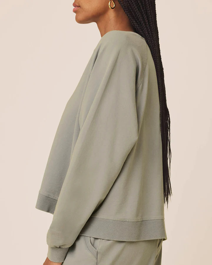 A side-profile view of a person wearing a light sage green cropped sweatshirt and matching sweatpants against a plain light beige studio background.
