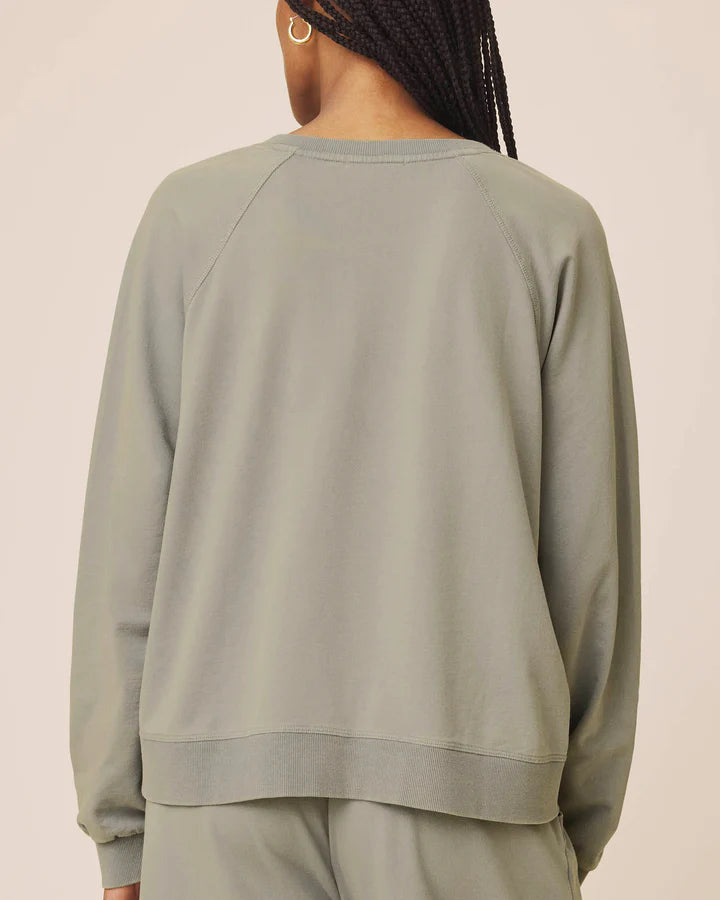 A person shown from the back wearing a plain crewneck sweatshirt and matching pants in a muted sage green color, against a solid light beige background.