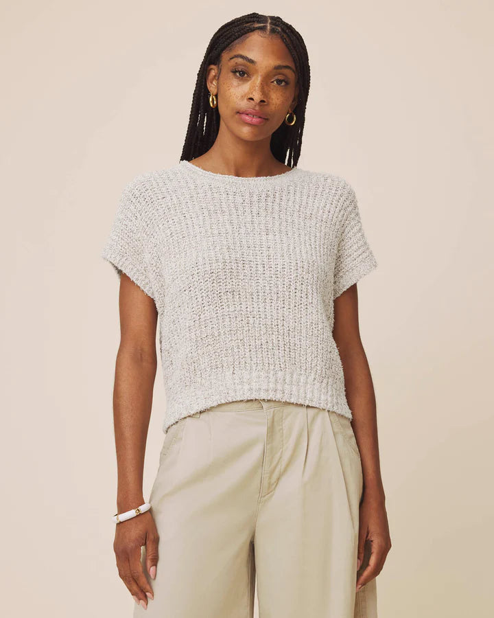 A woman modeling a short-sleeve, textured knit top in a light gray color, paired with light beige trousers, against a solid beige background.