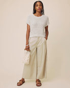 A woman modeling a light gray short-sleeve knit top, beige wide-leg trousers, brown sandals, and holding a white beaded handbag against a solid beige studio background.