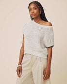 A woman modeling a textured knit off-the-shoulder short-sleeve top in a light gray color, paired with light beige trousers, against a solid beige background.