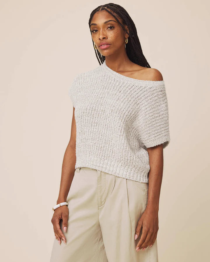 A woman modeling a textured knit off-the-shoulder short-sleeve top in a light gray color, paired with light beige trousers, against a solid beige background.