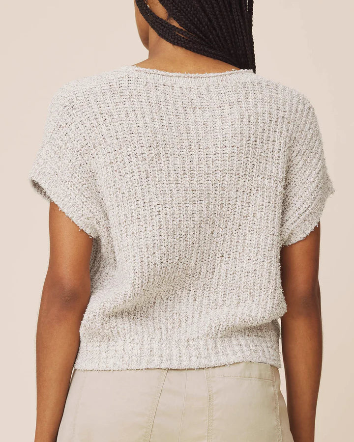 A person shown from the back wearing a short-sleeve textured knit top in a light heather gray color, paired with light beige pants, against a solid light beige background.