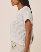A close-up side view of a woman wearing a textured, short-sleeve knit top in a light heather gray color, paired with beige pants, against a solid light beige background.