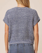 A person shown from the back wearing a short-sleeve textured knit top in a heathered blue-gray color, paired with blue jeans against a plain beige studio background.