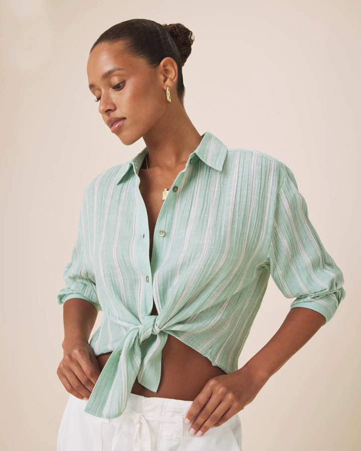 A woman wearing a mint green and white vertically striped button-down shirt tied at the waist, paired with white pants, against a plain beige background.