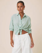 A woman modeling a mint green and white vertically striped button-down shirt tied at the waist, paired with white trousers, against a solid beige background.