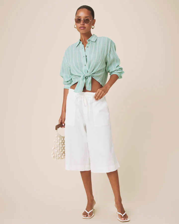 A woman modeling a mint green and white vertically striped button-down shirt tied at the waist, paired with white cropped pants, white sandals, and holding a small white beaded purse against a solid beige studio background.