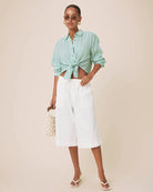 A woman modeling a mint green and white vertically striped button-down shirt tied at the waist, paired with white cropped pants, white sandals, and holding a small white beaded purse against a solid beige studio background.