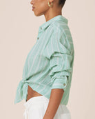 A side-profile view of a model wearing a mint green and white vertically striped cropped button-up shirt with rolled sleeves and a tied hem, paired with white pants against a solid pale beige background.
