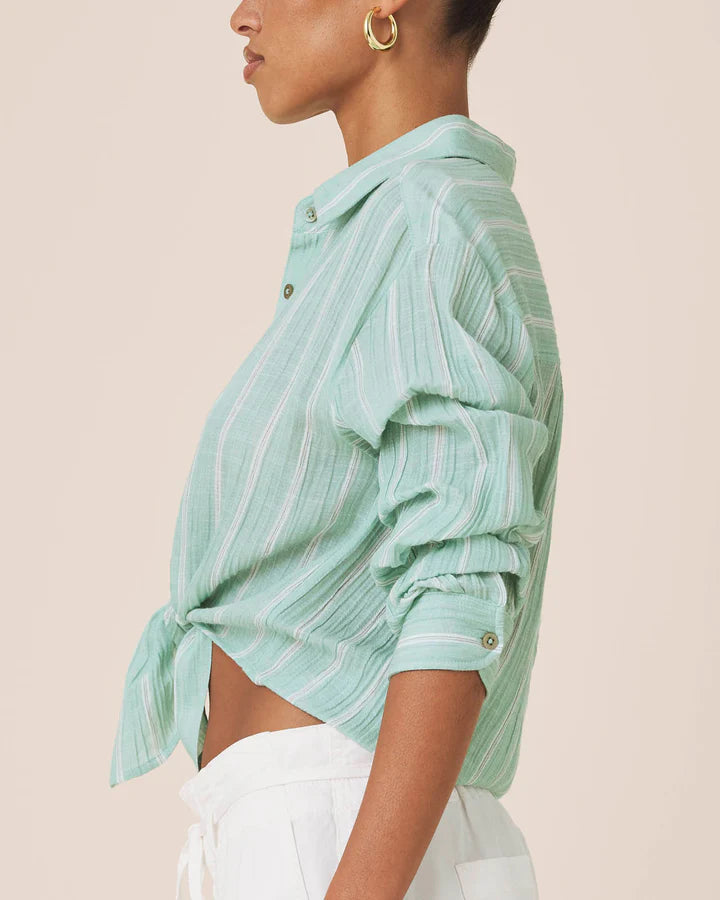 A side-profile view of a model wearing a mint green and white vertically striped cropped button-up shirt with rolled sleeves and a tied hem, paired with white pants against a solid pale beige background.