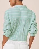 A woman shown from the back wearing a mint green and white striped long-sleeve blouse with a pleated back panel and white pants, against a plain beige background.