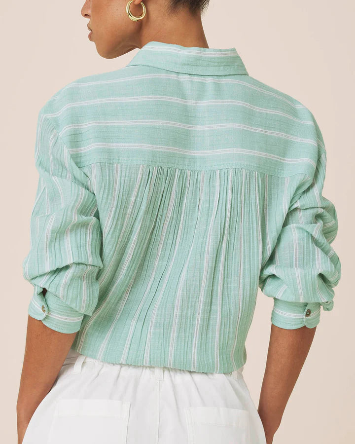 A woman shown from the back wearing a mint green and white striped long-sleeve blouse with a pleated back panel and white pants, against a plain beige background.