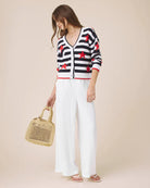 Woman wearing a black and white striped cardigan with red stars, white pants, and holding a straw bag on a beige background.