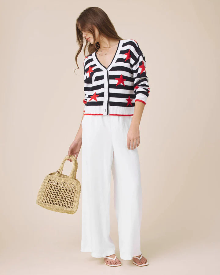 Woman wearing a black and white striped cardigan with red stars, white pants, and holding a straw bag on a beige background.