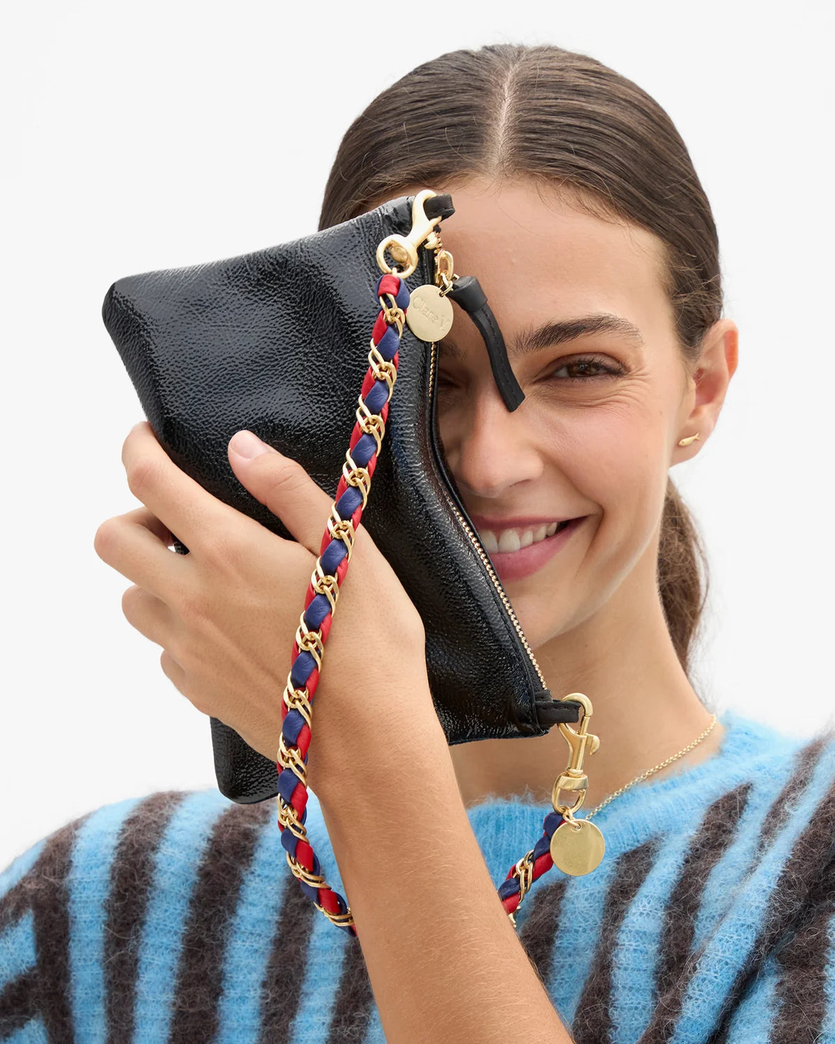 A woman holding a black textured leather clutch bag with a gold chain strap and red and blue braided accent details, set against a plain white background.