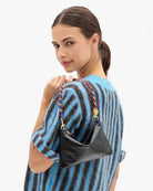 A woman wearing a blue and black striped knit sweater carries a small black leather shoulder bag with a braided red, blue, and gold chain strap against a plain white background.