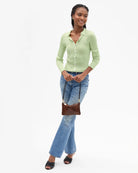 A woman modeling a light green ribbed knit button-up shirt, blue flare jeans, and black sandals, holding a small brown leather shoulder bag against a plain white studio background.