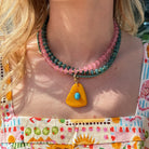 A close-up of a person wearing a double-strand beaded necklace with a yellow triangle pendant featuring a turquoise center. The necklace has one strand of pink beads and one strand of dark green beads.