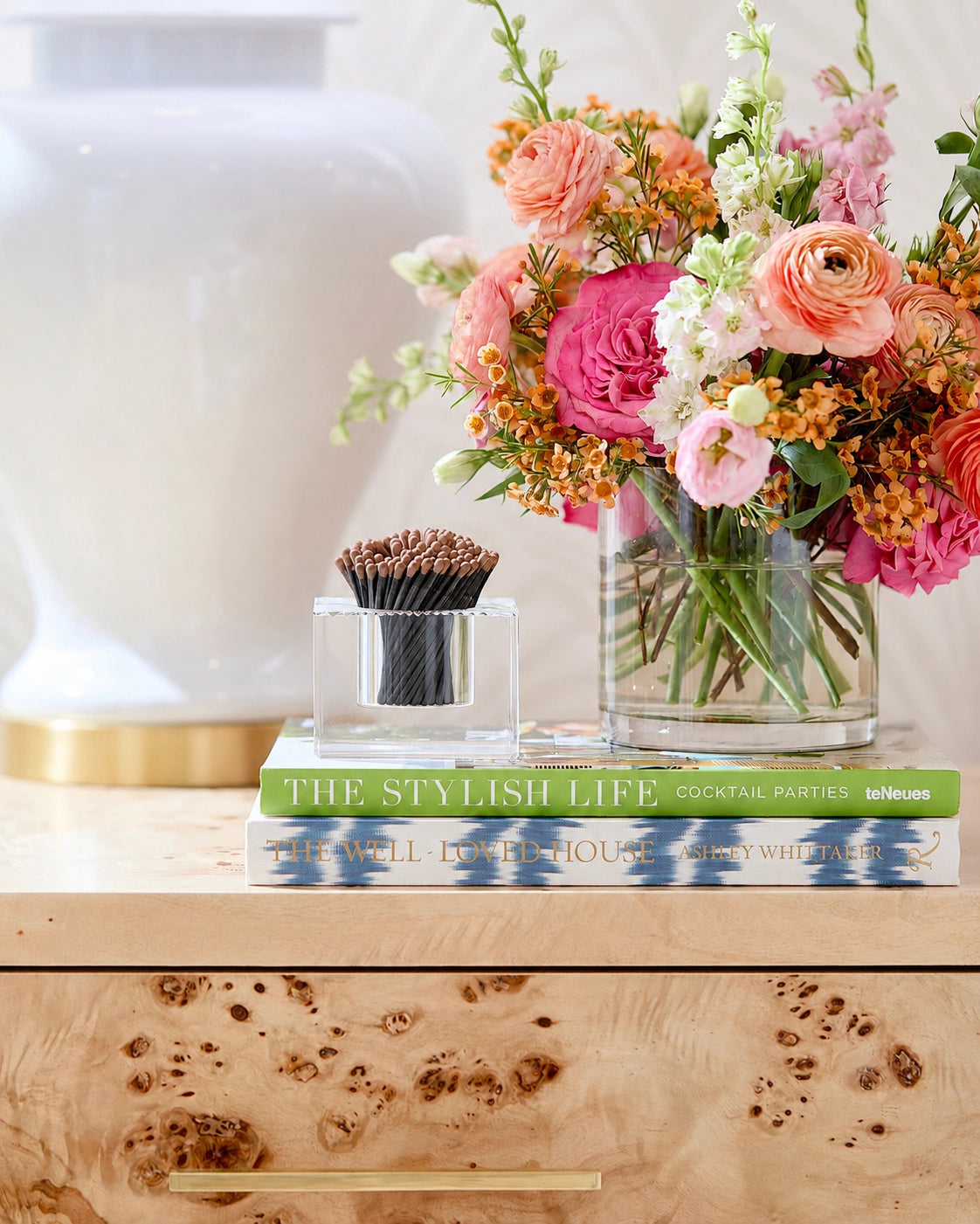 A styled interior scene showing a clear glass vase filled with a colorful floral arrangement on a light wood surface, alongside a small clear glass container holding a dark brown candle and a green book.
