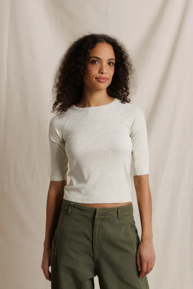 A woman with curly dark hair wearing a fitted short-sleeve knit top in off-white and olive green pants, standing against a draped cream fabric backdrop.
