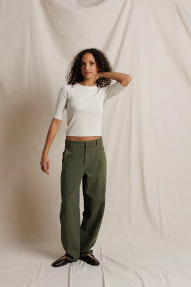 A model stands against a draped off-white fabric backdrop, wearing olive green wide-leg trousers, a white short-sleeve crop top, and black shoes.