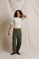 A model stands against a draped off-white fabric backdrop, wearing olive green wide-leg trousers, a white short-sleeve crop top, and black shoes.
