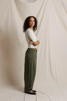 Woman standing against a plain white background wearing a white top and green pants.