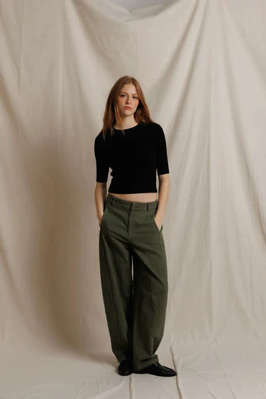 A model stands against a draped beige fabric backdrop, wearing a black short-sleeve cropped top and olive green wide-leg trousers.