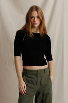 A woman modeling a black short-sleeve crop top paired with olive green trousers, standing against a draped beige fabric backdrop.