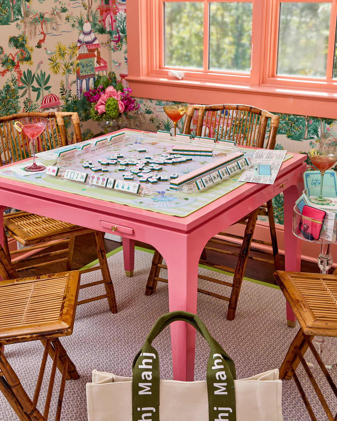 Pink dining table with a Mahjong game, surrounded by wooden chairs in a room with floral wallpaper.