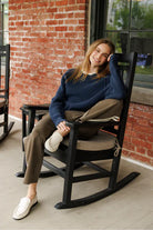 A woman sits in a black rocking chair on a porch, wearing a navy blue knit sweater, brown pants, and white slip-on shoes, with a red brick wall in the background.