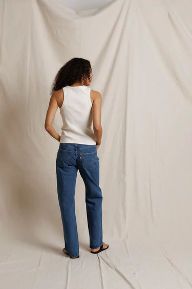 Person wearing a white sleeveless top and blue jeans standing against a plain background