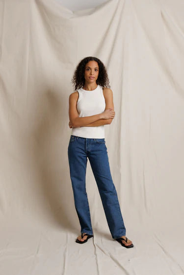 Woman wearing a white sleeveless top and blue jeans standing against a plain background