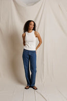 Woman wearing a white tank top and blue jeans standing against a plain background