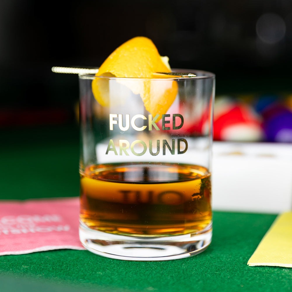 Glass of whiskey with a lemon slice on a green table, featuring the text 'FUCKED AROUND'.