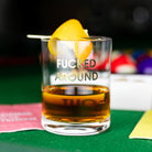 Glass of whiskey with a lemon slice on a green table, featuring the text 'FUCKED AROUND'.