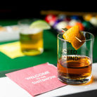 Glass of whiskey with a lemon garnish on a pool table with a pink napkin.
