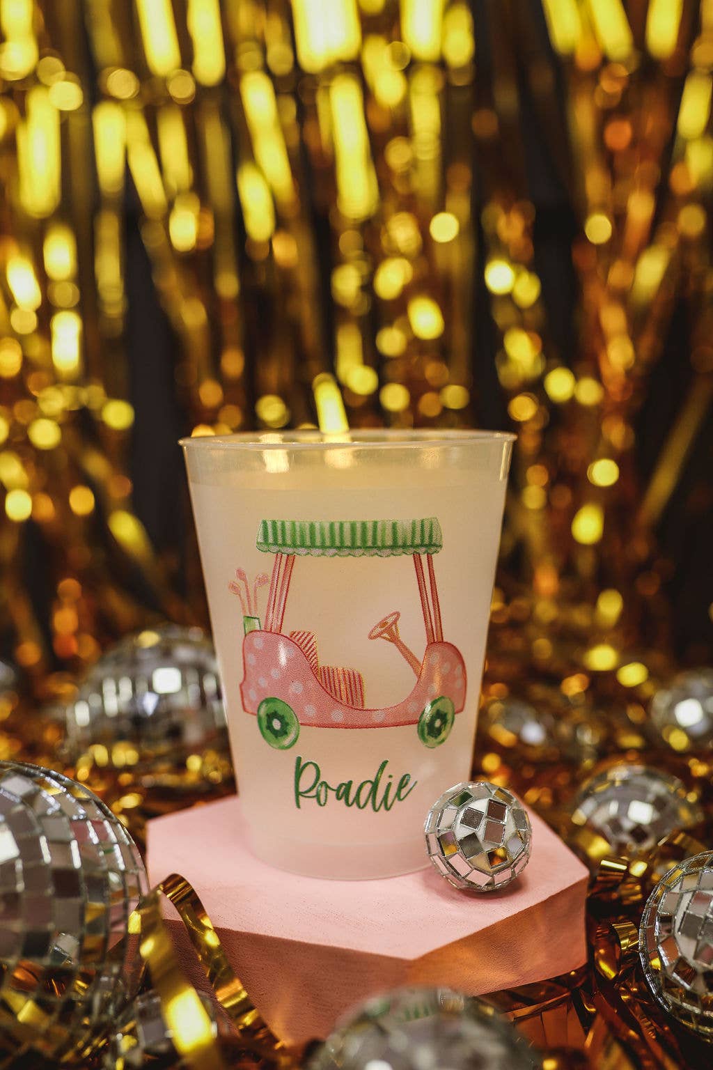 Candle in a pink holder with a design and text, placed on a reflective surface with a glittery gold background.