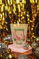 Candle in a pink holder with a design and text, placed on a reflective surface with a glittery gold background.