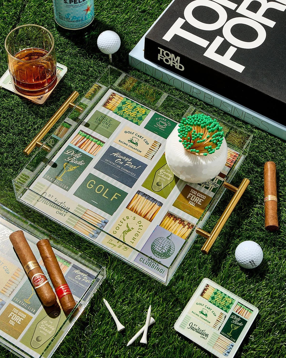 Golf-themed matchbox set with matches, a glass of whiskey, and golf balls on a grassy surface.