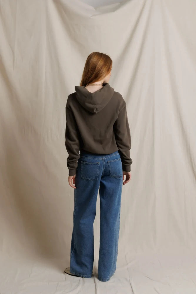 A person standing with their back to the camera, wearing a dark brown cropped hoodie and wide-leg blue jeans, posed in front of a draped beige fabric backdrop.
