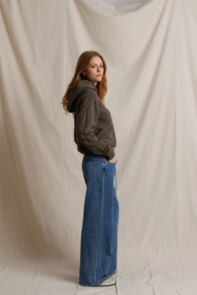 Person wearing a brown hoodie and blue jeans standing against a plain background