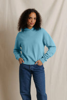 A woman with curly dark hair wearing a light blue long-sleeve mock-neck sweatshirt and blue jeans, standing against a draped beige fabric backdrop.