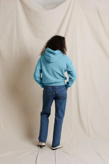 A person standing with their back to the camera, wearing a light blue hooded sweatshirt and blue jeans, posed against a draped off-white fabric backdrop.